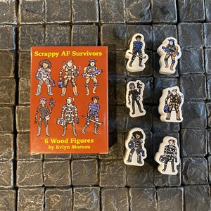 Puede incluir: Un conjunto de seis figuras de madera y una pequeña caja naranja. La caja dice "Scrappy AF Survivors" y presenta ilustraciones de las figuras. Las figuras están en varios atuendos y poses, con armas. Las figuras son de Evlyn Moreau.