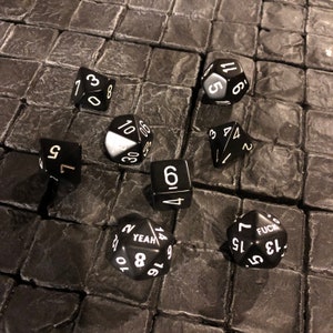 Puede incluir: Un conjunto de dados negros con números y texto blancos, incluyendo un D20 con "YEAH" y "FUCK". Los dados están dispersos sobre una superficie oscura y texturizada, ideal para juegos de mesa.