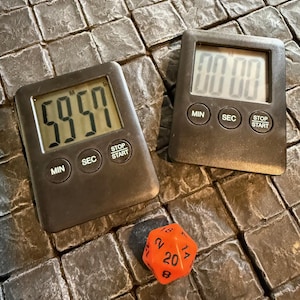 May include: Two black digital kitchen timers with a red 20-sided die. The timer on the left displays 59:57 and the timer on the right displays 00:00. Both timers have buttons labeled "MIN", "SEC", "STOP", and "START".