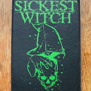 May include: Black rectangular patch with the words "Sickest Witch" in green, stylized lettering. Below the text is a green graphic of a skull and hands. The patch has a black border.