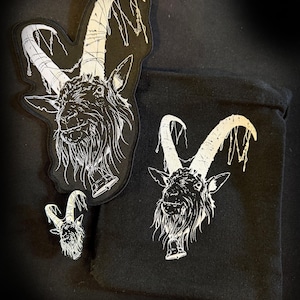 May include: Three black fabric patches with a white embroidered design of a goat's head with horns. The goat's head is facing forward with its mouth open. The horns are long and curved. The patches are different sizes. The largest patch is on the left, the medium patch is on the right, and the smallest patch is in the center.