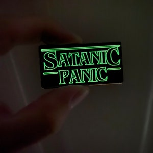 Könnte beinhalten: Eine schwarze Emaille-Anstecknadel mit einem grün leuchtenden Umriss. Die Nadel trägt die Aufschrift "SATANIC -PANIC-"