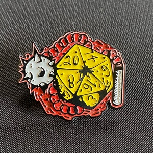 May include: A yellow 20-sided die with black numbers is surrounded by a red and white worm-like creature. The die is part of a pin with a silver backing.