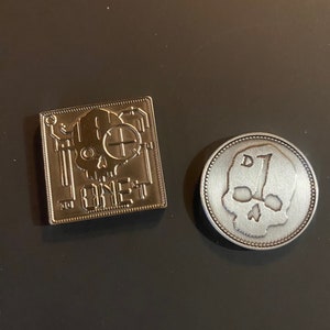 May include: Two silver metal tokens. One is square and features a skull with circuitry and the text "ONE-T". The other is round and features a skull with the number "1" above it.