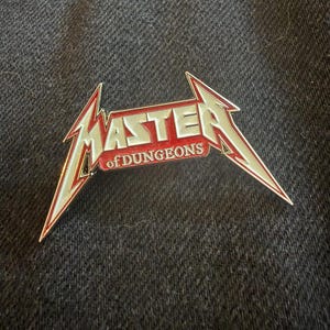 May include: A metal enamel pin featuring the word MASTER in a stylized, sharp-edged font with silver lettering and red accents. Below the word MASTER, the text of DUNGEONS is written in a smaller, white font against a red background.