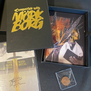 Könnte beinhalten: Ein schwarzes Box-Set mit einer goldenen Aufschrift, die "Kompatibel mit Mork Borg" lautet. Das Box-Set enthält ein Buch mit einem gold-schwarzen Design, eine schwarze Box mit einem gold-schwarzen Bild einer Person, die ein Buch hält, und eine kleine transparente Plastiktüte mit einer goldenen Münze darin.
