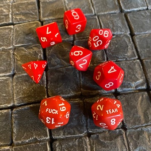 Puede incluir: Un conjunto de dados rojos con números blancos. Los dados están numerados del 1 al 6, 8, 10, 12, 14, 18, 19, 21 y 22. Un dado tiene la palabra "FUCK" y otro dado tiene la palabra "YEAH".