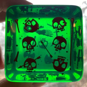 Può includere: Un vassoio quadrato in vetro verde con un design di teschi e ossa incrociate rosse. Il vassoio è riempito con un liquido trasparente e ha una finitura lucida.