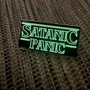 May include: A rectangular enamel pin with a black background and glowing green text that reads SATANIC PANIC. The lettering is stylized with horizontal lines extending from the top and bottom of the words, creating a bold, high-contrast design.