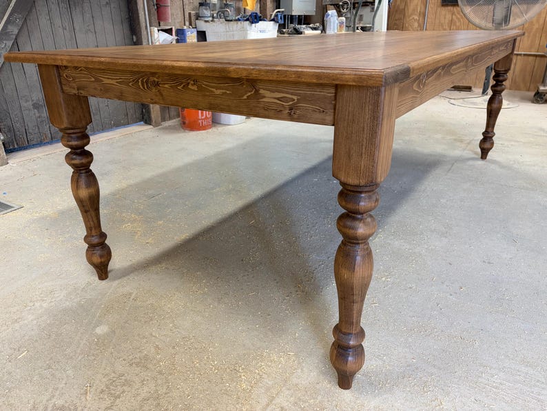 Peut inclure: Une table &agrave; manger rectangulaire en bois avec une finition teint&eacute;e brun moyen et un grain de bois visible. La table pr&eacute;sente une surface sup&eacute;rieure plate avec un bord droit et quatre pieds tourn&eacute;s avec un profil arrondi et d&eacute;coratif.