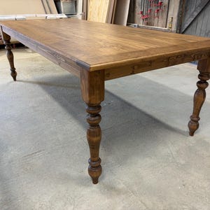 Peut inclure: Une table &agrave; manger rectangulaire en bois avec une finition teint&eacute;e brun moyen et une surface lisse et plate. La table pr&eacute;sente quatre pieds &eacute;pais et tourn&eacute;s avec des d&eacute;tails arrondis d&eacute;coratifs, offrant une base solide au grand plateau.