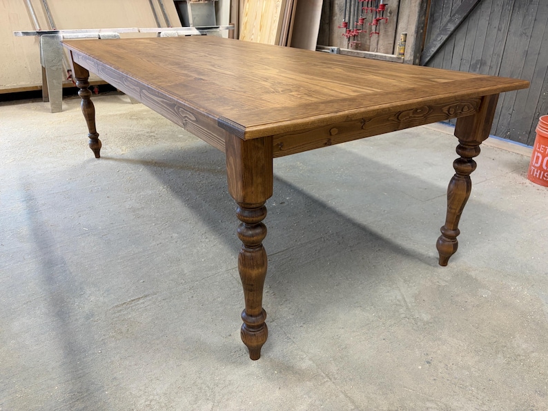 Peut inclure: Une table &agrave; manger rectangulaire en bois avec une finition teint&eacute;e brun moyen et une surface lisse et plate. La table pr&eacute;sente quatre pieds &eacute;pais et tourn&eacute;s avec des d&eacute;tails arrondis d&eacute;coratifs, offrant une base solide au grand plateau.