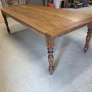 Peut inclure: Une table &agrave; manger rectangulaire en bois avec une finition teint&eacute;e brun moyen et un grain de bois visible. La table pr&eacute;sente quatre pieds &eacute;pais et tourn&eacute;s avec un profil d&eacute;coratif arrondi et un plateau plat et lisse aux bords droits.