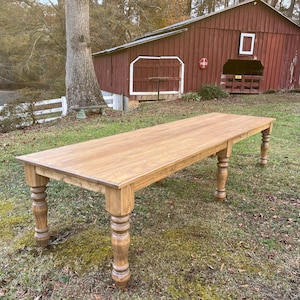 May include: A long, rectangular wooden table with a light brown finish. The table has four turned legs and is set outdoors on a grassy area. The background includes a red barn and a white fence.