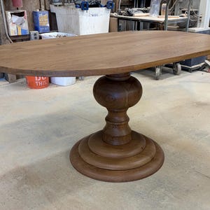 May include: An oval dining table crafted from medium-brown wood with a visible grain pattern. The table features a smooth, flat top surface supported by a single, thick, central pedestal base that rests on a wide, tiered circular foundation.
