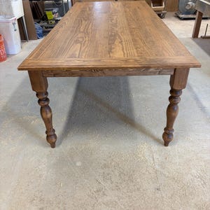 Peut inclure: Une table &agrave; manger rectangulaire en bois avec une finition teint&eacute;e brun moyen et un grain de bois visible. La table pr&eacute;sente un plateau plat avec un cadre de bordure et quatre pieds tourn&eacute;s de style fuseau qui s'affinent vers le bas.