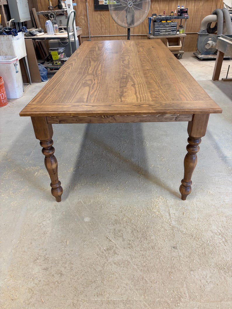 Peut inclure: Une table &agrave; manger rectangulaire en bois avec une finition teint&eacute;e brun moyen et un grain de bois visible. La table pr&eacute;sente un plateau plat avec un cadre de bordure et quatre pieds tourn&eacute;s de style fuseau qui s'affinent vers le bas.