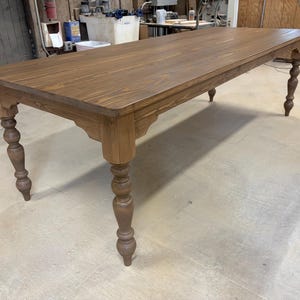 May include: A large, rectangular wooden table with a warm brown finish. The table features ornate, turned legs and decorative detailing along the apron. The table's dimensions are approximately 96 inches long and 36 inches wide.