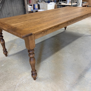 Peut inclure: Une table &agrave; manger rectangulaire en bois avec une finition teint&eacute;e brun moyen et un grain de bois visible. La table pr&eacute;sente quatre pieds tourn&eacute;s avec un design arrondi et bulbeux, ainsi qu'une surface de plateau lisse et plate.