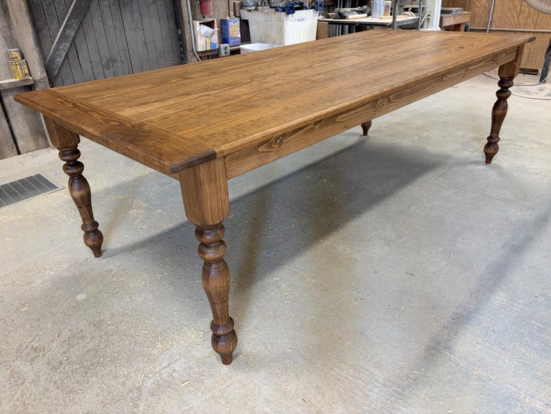 Peut inclure: Une table &agrave; manger rectangulaire en bois avec une finition teint&eacute;e brun moyen et un grain de bois visible. La table pr&eacute;sente quatre pieds tourn&eacute;s avec un design arrondi et bulbeux, ainsi qu'une surface de plateau lisse et plate.