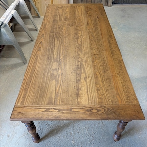 Peut inclure: Une table &agrave; manger rectangulaire en bois avec une finition teint&eacute;e brun moyen et des motifs de grain de bois visibles. La table pr&eacute;sente une surface sup&eacute;rieure plate avec un cadre et quatre pieds tourn&eacute;s, vue en plong&eacute;e.