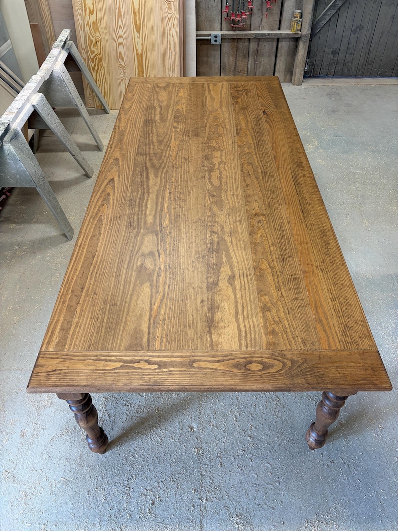Peut inclure: Une table &agrave; manger rectangulaire en bois avec une finition teint&eacute;e brun moyen et des motifs de grain de bois visibles. La table pr&eacute;sente une surface sup&eacute;rieure plate avec un cadre et quatre pieds tourn&eacute;s, vue en plong&eacute;e.