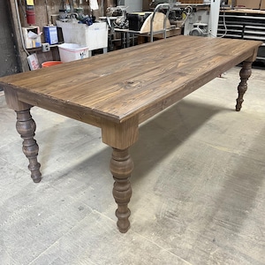 May include: A large, rustic wooden table with a dark brown stain. The table has a rectangular top and four turned legs. The legs are made of wood and have a decorative design.