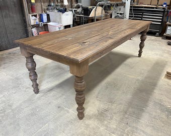 May include: A large, rustic wooden table with a dark brown stain. The table has a rectangular top and four turned legs. The legs are made of wood and have a decorative design.