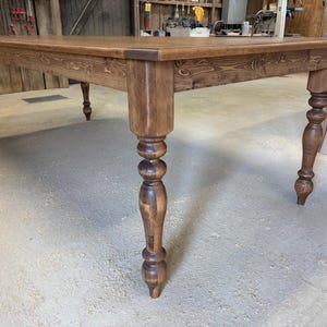 Peut inclure: Une grande table &agrave; manger rectangulaire en bois avec une finition teint&eacute;e brun riche et des motifs de grain de bois visibles. La table est soutenue par quatre pieds &eacute;pais et tourn&eacute;s au design classique, montrant une construction robuste.