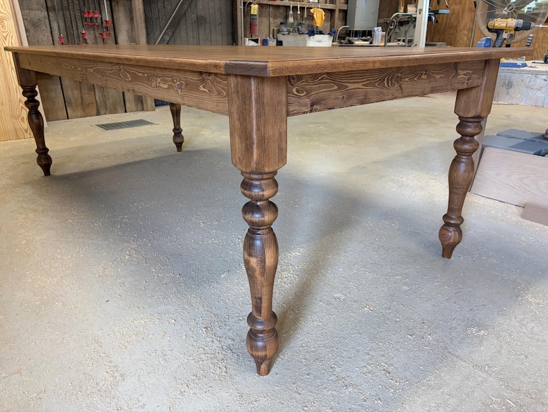 Peut inclure: Une grande table &agrave; manger rectangulaire en bois avec une finition teint&eacute;e brun riche et des motifs de grain de bois visibles. La table est soutenue par quatre pieds &eacute;pais et tourn&eacute;s au design classique, montrant une construction robuste.