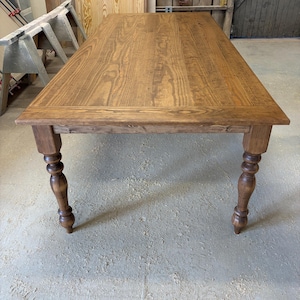 Peut inclure: Une table &agrave; manger rectangulaire en bois avec une finition teint&eacute;e brun moyen et un grain de bois visible. La table pr&eacute;sente un plateau plat avec un cadre de bordure et quatre pieds tourn&eacute;s avec des d&eacute;tails arrondis et bomb&eacute;s.