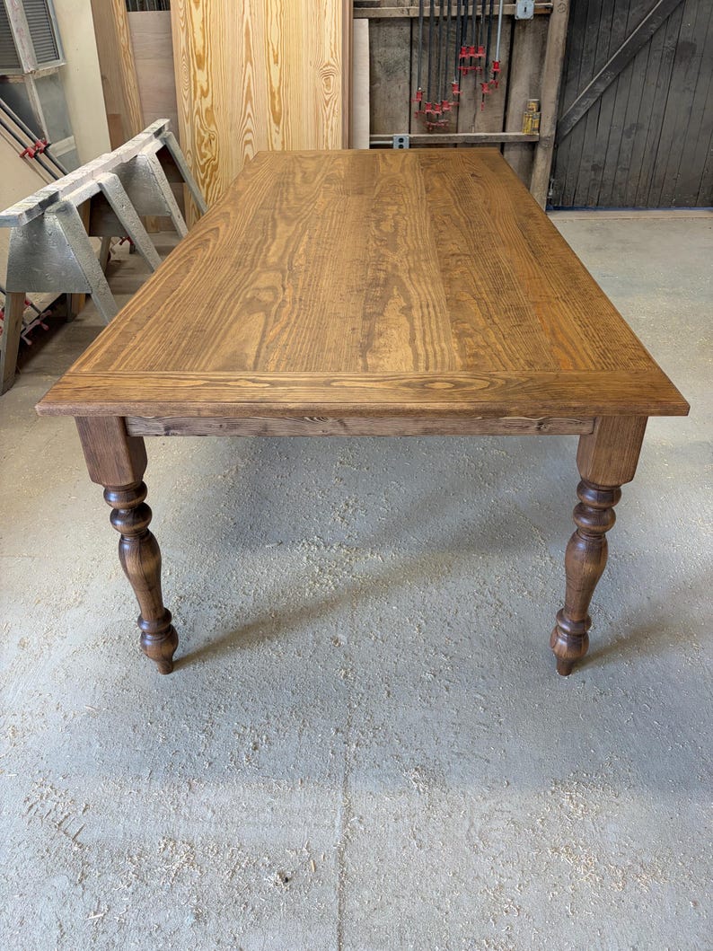 Peut inclure: Une table &agrave; manger rectangulaire en bois avec une finition teint&eacute;e brun moyen et un grain de bois visible. La table pr&eacute;sente un plateau plat avec un cadre de bordure et quatre pieds tourn&eacute;s avec des d&eacute;tails arrondis et bomb&eacute;s.