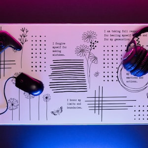 以下が含まれることがあります： 白地に黒と白のイラストが描かれたデスクマット。花、蝶、幾何学模様が描かれている。マットには「私は自分の過ちを許します」「私は自分の世代のために癒されるために全責任を負います」「私は自分の限界と境界線を尊重します」「私の感情と行動」というテキストが書かれている。