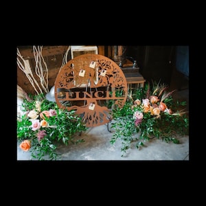 May include: A copper-colored metal tree with the word "Finch" cut out of the trunk. The tree is surrounded by greenery and pink flowers.