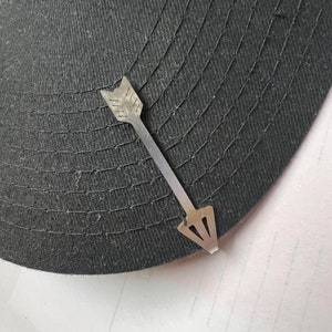 May include: A silver arrow-shaped hat clip. The clip is attached to a black baseball cap.