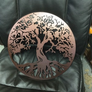 May include: A copper-colored metal tree of life wall decor with a circular shape and intricate details. The tree has a thick trunk and branches with leaves, and the roots are spread out at the bottom.