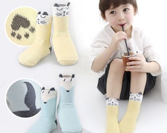 May include: A pair of light yellow and a pair of light blue children's socks. The yellow socks have a cat face design at the top and paw prints on the bottom. The blue socks have a fox face design at the top.
