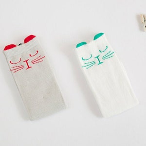 May include: Two pairs of white knee-high socks with cat faces. One pair has red cat ears and red whiskers, the other has green cat ears and green whiskers.