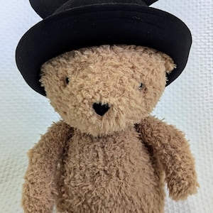 May include: A plush teddy bear with light brown fur, wearing a black hat with attached ears. The bear has a heart-shaped nose and is sitting upright. The background is a white, textured surface.