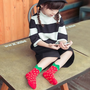 May include: A young person wearing a black and white striped top and black shorts, paired with red socks featuring a strawberry design. The socks have green cuffs and yellow seeds. The person is holding a smartphone.