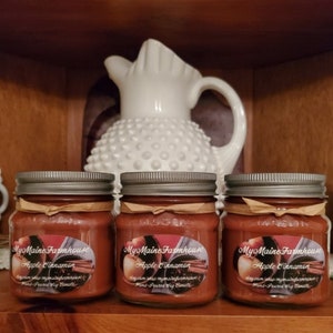 May include: Three glass jars with silver lids filled with brown candle wax. Each jar has a label that reads "My Maine Farmhouse Apple Cinnamon Hand-Poured Soy Candle".