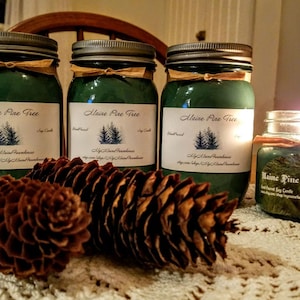 May include: Three green glass jars with silver lids and labels that read "Maine Pine Tree" soy candles. The jars are sitting on a white lace tablecloth with pine cones in front of them. One jar is lit and the flame is visible.