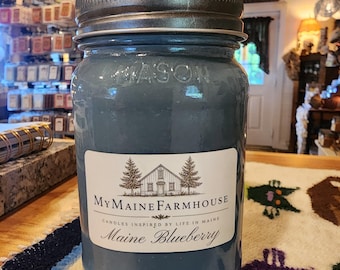 May include: A blue candle in a glass mason jar with a dark metal lid. The white label features a black line drawing of a farmhouse between two pine trees, with the text MyMaineFarmhouse, Candles Inspired by Life in Maine, and Maine Blueberry.