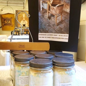 May include: A collection of glass jars filled with white laundry soap flakes. The jars have black lids and are labeled "Clean Laundry" with instructions to use 1-3 tablespoons per load. The jars are sitting on a white surface. A sign above the jars reads "Handmade Laundry Soap - 16 oz./$9.99 Use one to three tablespoons per load."