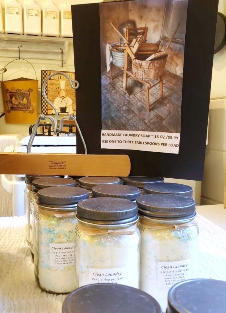 May include: A collection of glass jars filled with white laundry soap flakes. The jars have black lids and are labeled "Clean Laundry" with instructions to use 1-3 tablespoons per load. The jars are sitting on a white surface. A sign above the jars reads "Handmade Laundry Soap - 16 oz./$9.99 Use one to three tablespoons per load."