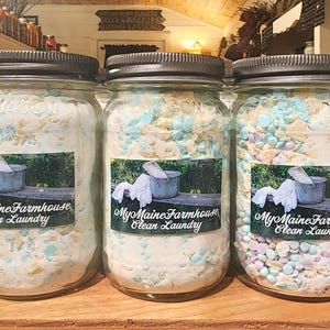 Puede incluir: Tres frascos de vidrio llenos de detergente para la ropa. Los frascos tienen tapas negras y etiquetas que dicen "My Maine Farmhouse Clean Laundry".