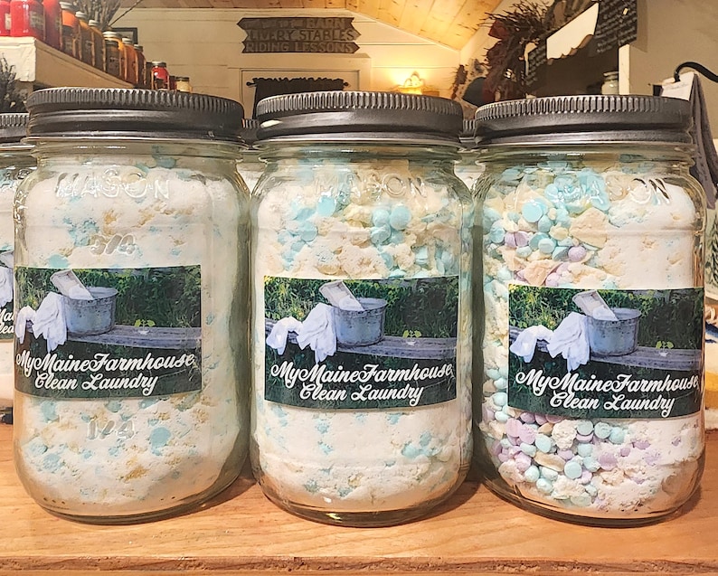 May include: Three glass jars filled with laundry detergent. The jars have black lids and labels that say "My Maine Farmhouse Clean Laundry".