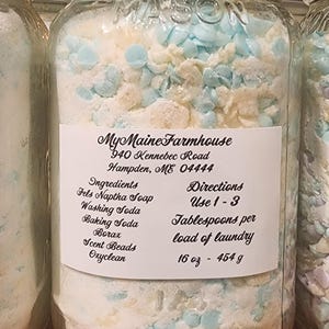 May include: A glass jar filled with white laundry detergent and blue scent beads. The label reads "My Maine Farmhouse", "940 Kennebec Road", "Hampden, ME 04444", "Ingredients: Fels Naptha Soap, Washing Soda, Baking Soda, Borax, Scent Beads, Oxyclean", "Directions: Use 1-3 Tablespoons per load of laundry", "16 oz - 454 g".
