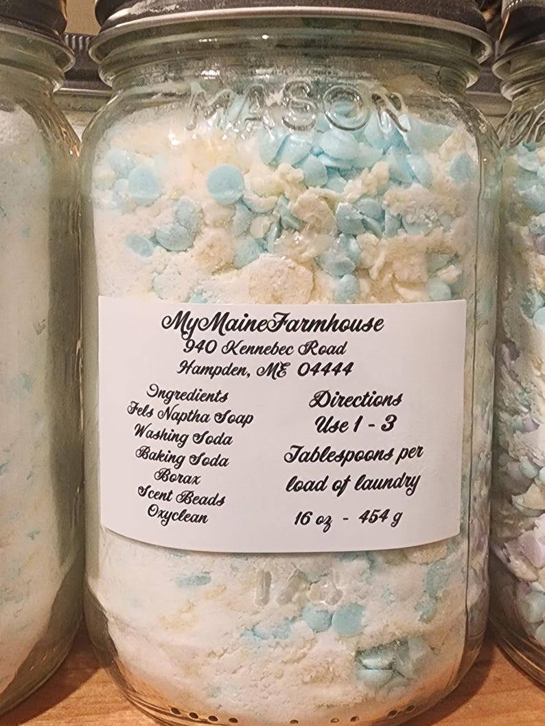 May include: A glass jar filled with white laundry detergent and blue scent beads. The label reads "My Maine Farmhouse", "940 Kennebec Road", "Hampden, ME 04444", "Ingredients: Fels Naptha Soap, Washing Soda, Baking Soda, Borax, Scent Beads, Oxyclean", "Directions: Use 1-3 Tablespoons per load of laundry", "16 oz - 454 g".