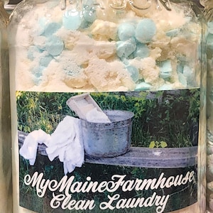 May include: A glass jar filled with white laundry detergent and blue beads. The jar has a black lid and a label that reads "My Maine Farmhouse Clean Laundry".